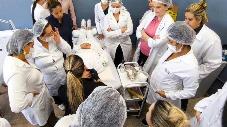 Estética em 3 dimensões: participe da aula gratuita e eleve seus resultados 8 Grupo de alunas em aula prática de estética, reunidas ao redor de uma paciente deitada enquanto uma professora demonstra técnicas com equipamentos clínicos.