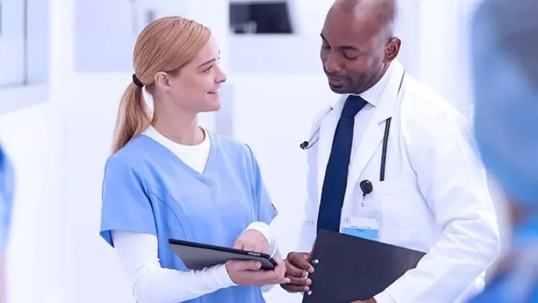 Dois profissionais da saúde conversando em um corredor clínico, um segurando um tablet e outro com estetoscópio no pescoço, simbolizando integração entre áreas assistenciais e administrativas.
