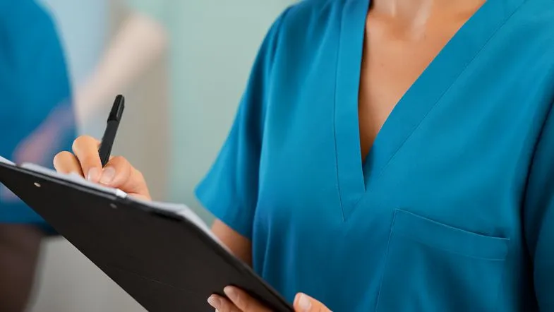 Pós-graduação em Gestão e Metodologia ONA: liderando com excelência 9 Profissional da saúde escrevendo em prancheta, sem estetoscópio, enfatizando organização, registro clínico e atuação cuidadosa em ambiente hospitalar.