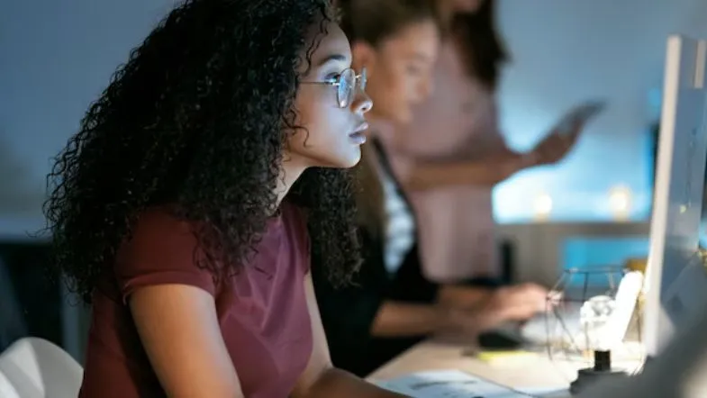 Faculdade ITH realiza masterclass gratuita sobre produtividade, foco e resultados reais 9 Mulher jovem trabalhando concentrada no computador à noite em ambiente de escritório moderno.