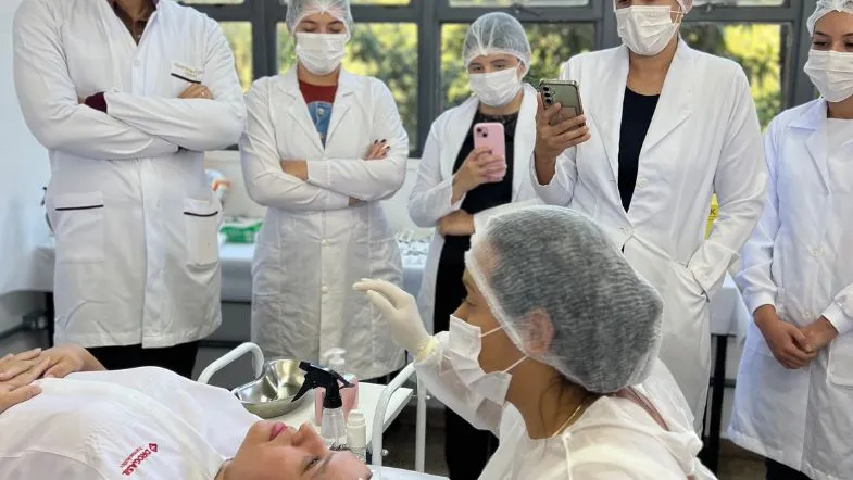 Estética em 3 dimensões: participe da aula gratuita e eleve seus resultados 9 Sala de aula com profissionais de jaleco observando demonstração prática de procedimento estético, enquanto uma instrutora realiza a técnica em uma paciente deitada.