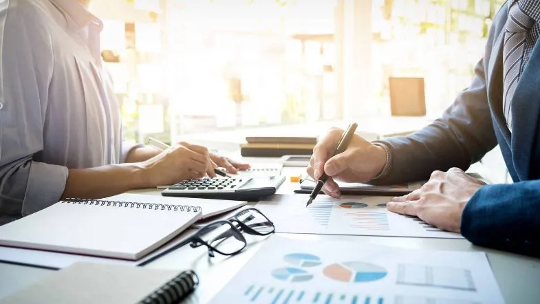 Qual é a diferença entre Administração e Ciências Contábeis? Saiba qual curso escolher! 6 Duas pessoas trabalhando em conjunto, analisando gráficos e utilizando calculadora e papéis com planilhas sobre uma mesa iluminada.
