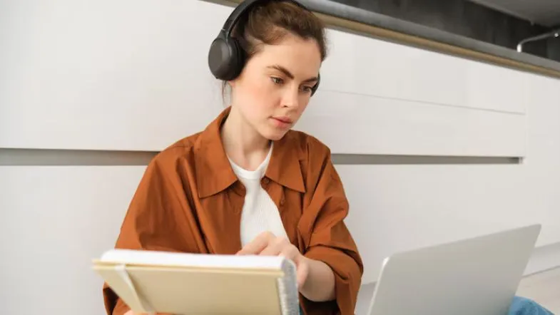Nova política do MEC vai afetar minha graduação EAD? Entenda o que muda 6 Jovem estudante com fones de ouvido e caderno nas mãos, concentrada em uma aula online no laptop.
