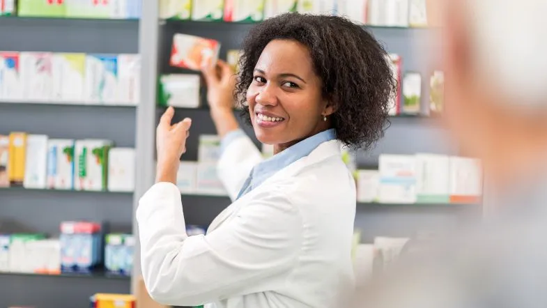 Farmacêutica sorridente em atendimento na farmácia clínica, organizando prateleiras com medicamentos.