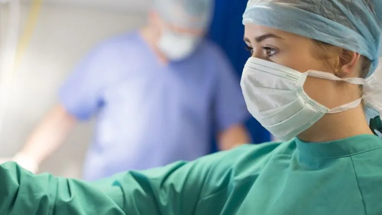 O que faz um enfermeiro de UTI? Veja as funções e competências essenciais 7 Profissional de saúde com máscara e touca concentrada em um procedimento dentro do ambiente hospitalar.