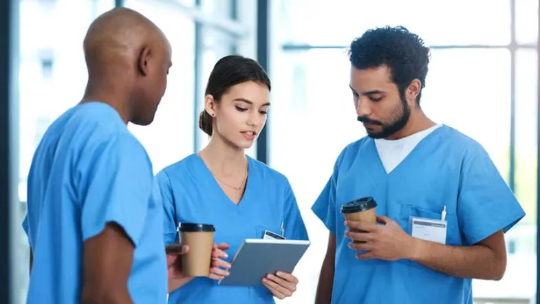 Quais áreas de pós-graduação em Enfermagem estão em alta? Confira as mais requisitadas 7 Enfermeira em uniforme azul digitando no notebook em um consultório, com cadeira de rodas e maca ao fundo.