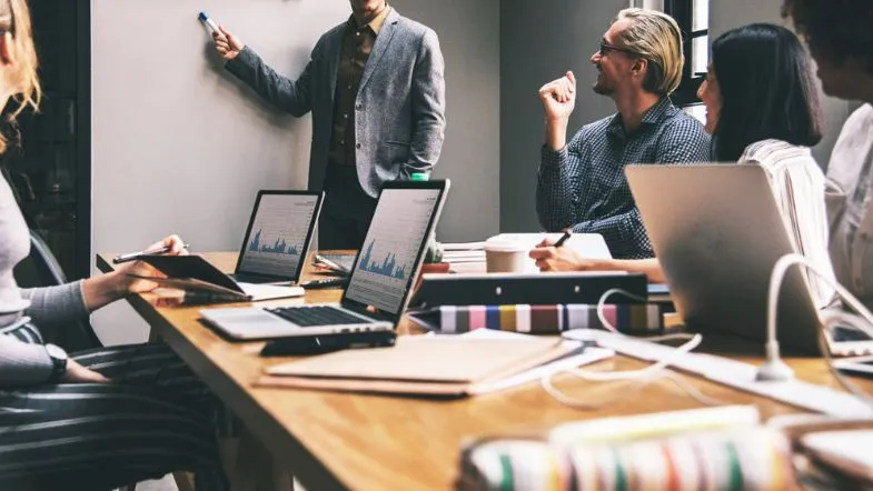 Qual é a diferença entre Administração e Ciências Contábeis? Saiba qual curso escolher! 7 Grupo de profissionais reunidos em uma sala de reunião, com laptops abertos e um homem apresentando algo em um quadro branco.