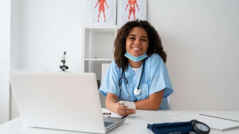 Enfermeira sorridente com jaleco e notebook na mesa, simbolizando oportunidades de qualificação por meio de pós-graduação EAD em saúde.