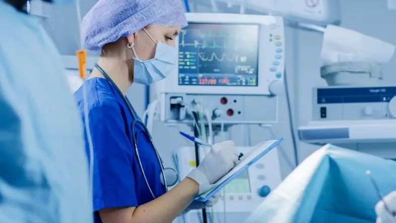 Técnica em enfermagem usando máscara e avental enquanto registra informações de paciente em unidade de terapia intensiva.