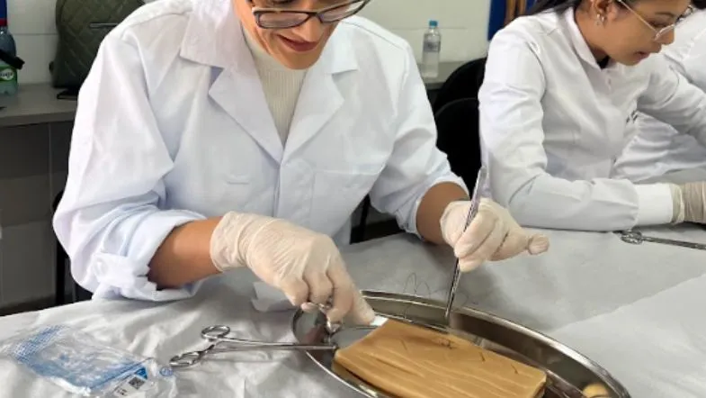 Enfermeira em treinamento prático de sutura obstétrica, manipulando instrumentos cirúrgicos em bancada de simulação.
