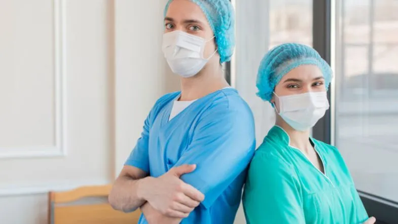 O que faz um enfermeiro de UTI? Veja as funções e competências essenciais 8 Enfermeiro de UTI e enfermeira de UTI vestidos com aventais cirúrgicos e máscaras, posando no corredor do hospital com postura confiante.