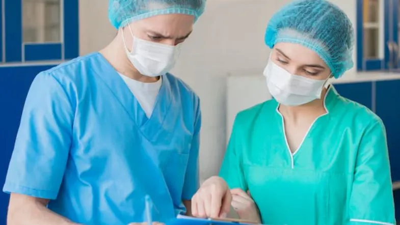 Quais áreas de pós-graduação em Enfermagem estão em alta? Confira as mais requisitadas 8 Três profissionais de enfermagem em uniforme azul conversando enquanto seguram copos de café e um tablet, demonstrando troca de informações no trabalho.