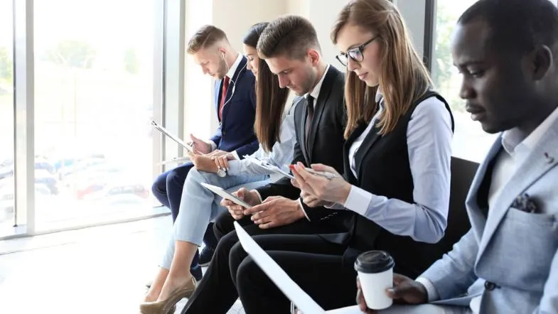 Nova política do MEC vai afetar minha graduação EAD? Entenda o que muda 8 Grupo de jovens profissionais aguardando uma entrevista, com roupas formais e celulares em mãos.