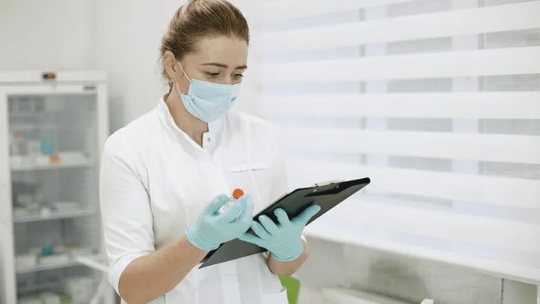 Farmacêutica com EPI avaliando medicamento e registrando dados em ambiente clínico.
