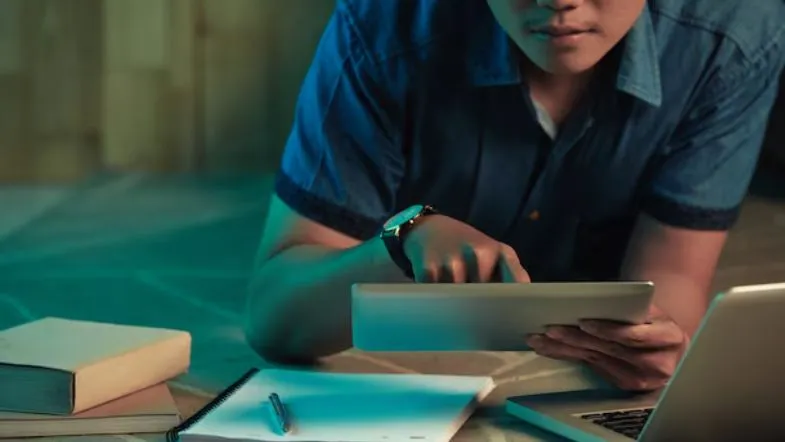 Edtechs: o que são e por que estão transformando a educação? 8 Jovem estudando com tablet, laptop e caderno sobre a mesa, em ambiente moderno e escuro, representando a liberdade oferecida pelas edtechs de ensino.