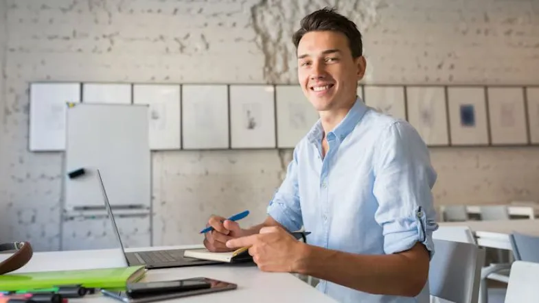 Qual é a diferença entre Administração e Ciências Contábeis? Saiba qual curso escolher! 9 Jovem sorridente sentado em uma mesa com notebook, caderno e caneta, em um ambiente de trabalho moderno com quadro branco ao fundo, representando o futuro de quem cursa Administração.
