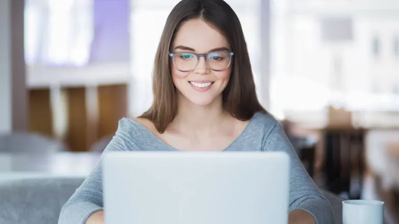 Mulher estudando em casa no computador com sorriso, exemplificando a praticidade e a flexibilidade dos cursos de pós-graduação EAD em saúde.