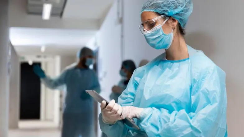 Formação profissional no combate às hepatites virais: por que é essencial? 9 Profissional de saúde utilizando equipamentos de proteção individual (EPI) e consultando um tablet em ambiente hospitalar, demonstrando atenção à biossegurança.
