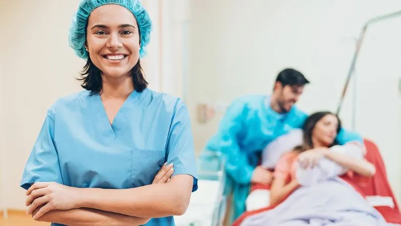 Atuação da enfermeira obstetra no parto humanizado: o que você precisa saber 9 Enfermeira sorridente em primeiro plano, enquanto ao fundo um casal celebra o nascimento do bebê na sala de parto.