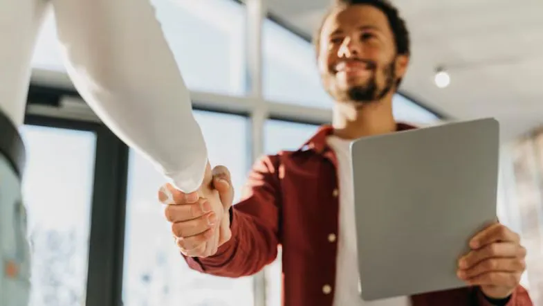 Nova política do MEC vai afetar minha graduação EAD? Entenda o que muda 9 Homem sorridente apertando a mão de outro profissional, segurando um tablet, em ambiente corporativo.