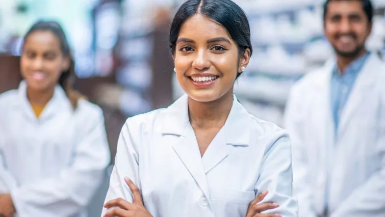 Jovem farmacêutica sorridente em consultório farmacêutico com outros colegas ao fundo.
