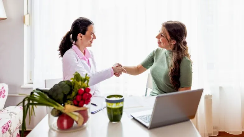 Profissional da Nutrição cumprimenta paciente sorridente ao final da consulta em consultório com frutas e notebook sobre a mesa.