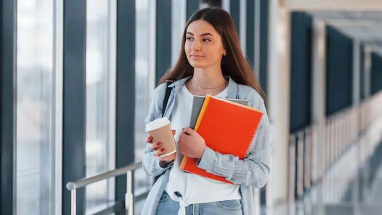 Como se preparar para o ENEM 2025 e conquistar sua vaga na faculdade 9 Estudante universitária segurando café e cadernos em corredor da faculdade.