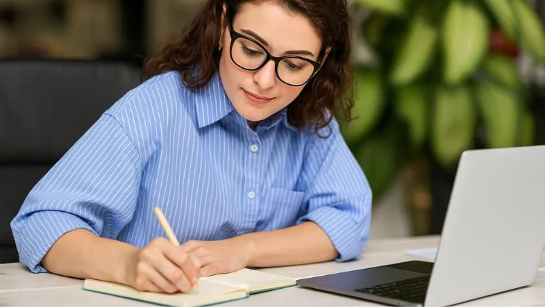 Edtechs: o que são e por que estão transformando a educação? 9 Mulher escrevendo em caderno ao lado de um notebook, concentrada em ambiente com plantas ao fundo.