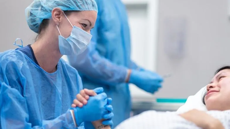 Enfermeira obstétrica prestando apoio emocional a gestante durante o trabalho de parto, com ambiente hospitalar ao fundo.