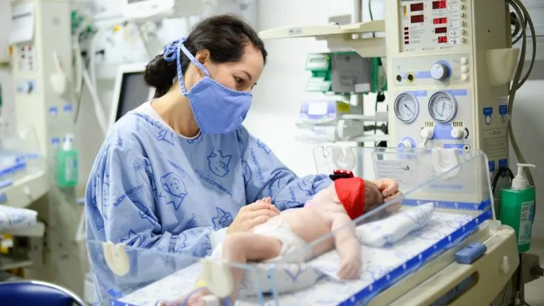 Enfermeira cuidando de recém-nascido em incubadora com equipamentos hospitalares na UTI Neonatal.