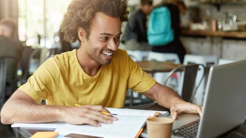 Rapaz sorridente com camiseta amarela estudando em um café, com livros abertos e notebook à sua frente.