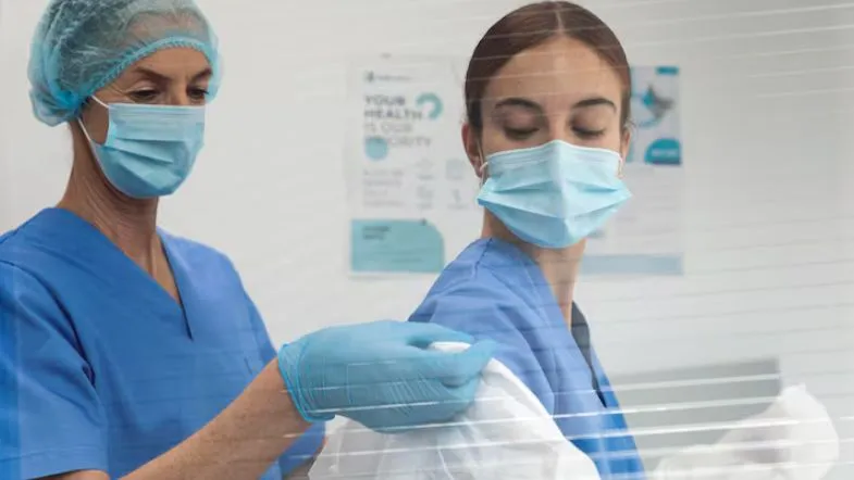 Profissional de saúde auxiliando colega na colocação de avental, ambas usando máscaras e toucas, dentro de clínica ou hospital, enfatizando a importância do SCIRAS.