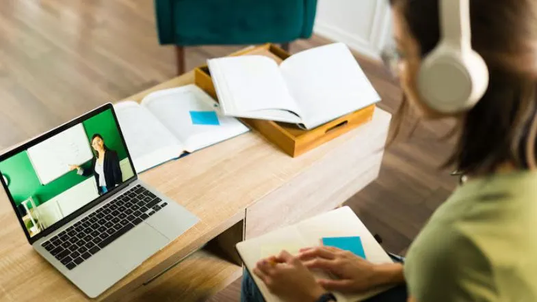 Novo marco regulatório da Educação a Distância: o que muda e quem será impactado 6 Aluna assistindo a uma aula online ao vivo pelo notebook, usando fones de ouvido e fazendo anotações com livros abertos.