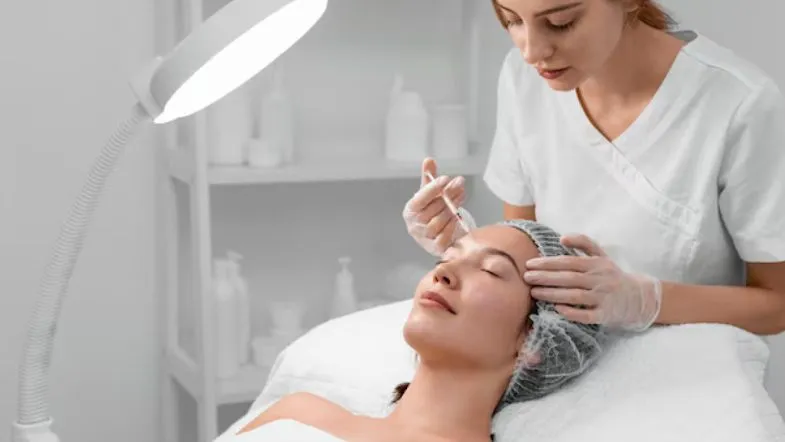 Estética de Alta Performance: formação completa em botox, preenchimento e laserterapia 6 steta loira aplicando injetável em paciente deitada em clínica estética, sob iluminação direta.