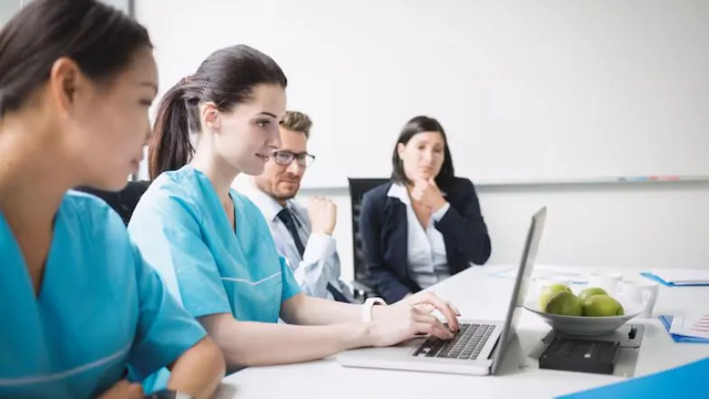 10 caminhos na pós-graduação para transformar sua carreira em Enfermagem 6 Grupo de enfermeiras e profissionais de saúde participando de uma reunião de capacitação, com foco em uma enfermeira usando laptop.