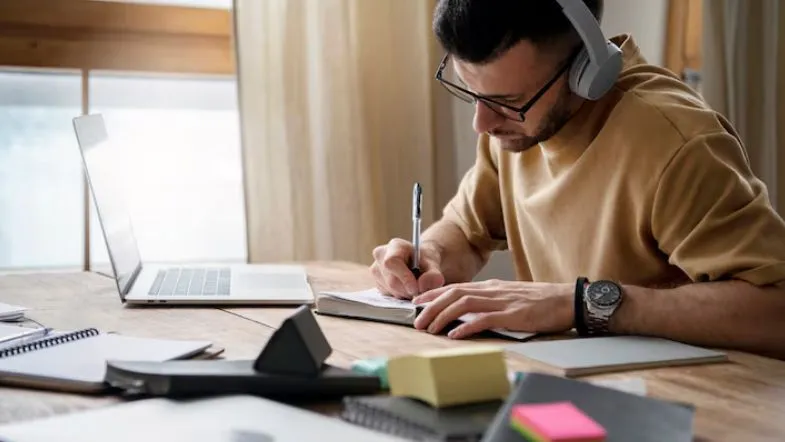 Novo marco regulatório da Educação a Distância: o que muda e quem será impactado 7 Homem estudando na modalidade de educação a distância com fones de ouvido, escrevendo em caderno ao lado de notebook e materiais escolares.