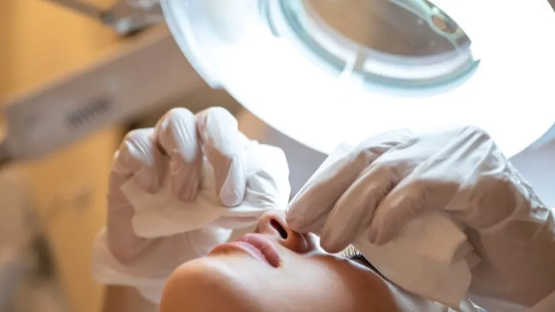 Estética de Alta Performance: formação completa em botox, preenchimento e laserterapia 7 Profissional da estética realizando procedimento com luvas e luz focal no rosto da paciente.
