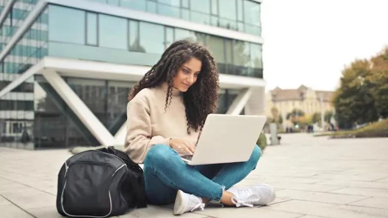Como escolher o curso de graduação ideal para você: dicas práticas para decidir com segurança 7 Estudante universitária utilizando notebook em frente ao prédio moderno, destacando a flexibilidade da graduação EAD.