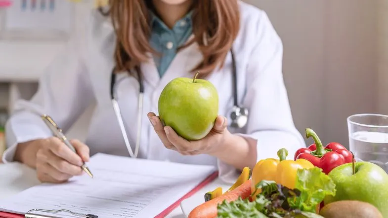 Nutricionista escreve em prontuário enquanto segura maçã verde, ao lado de mesa com frutas e vegetais coloridos.