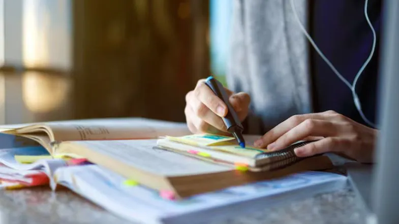 Pessoa destacando informações em um caderno com marca-texto, ao lado de livros e post-its.