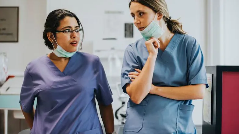 Duas enfermeiras conversando em corredor hospitalar, com máscaras abaixadas no pescoço.