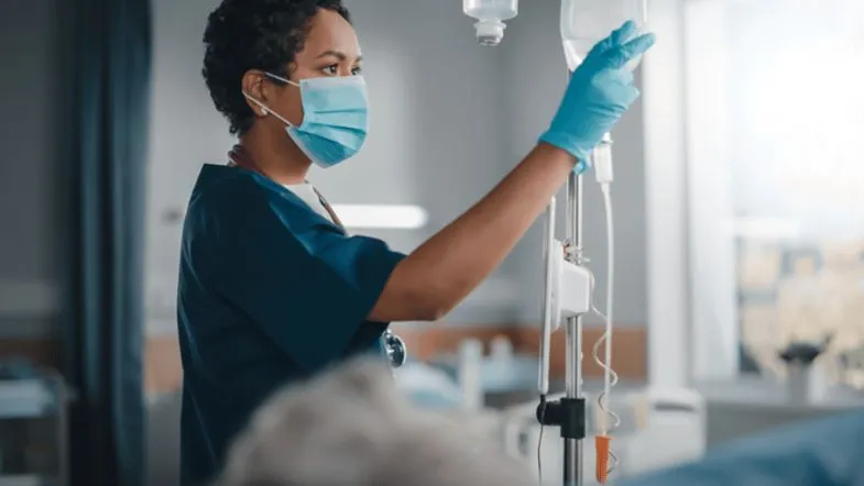 10 caminhos na pós-graduação para transformar sua carreira em Enfermagem 8 Profissional da Enfermagem ajustando soro fisiológico em paciente, usando máscara e luvas, em ambiente hospitalar.