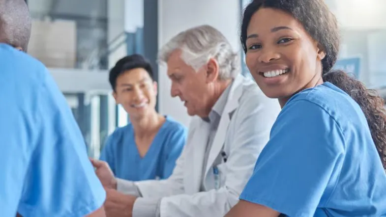 Grupo de profissionais da saúde reunidos, incluindo médicos e enfermeiros, com destaque para uma enfermeira sorrindo em primeiro plano.