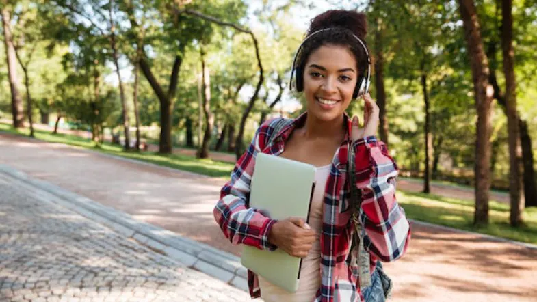 Novo marco regulatório da Educação a Distância: o que muda e quem será impactado 9 Jovem estudante sorridente com notebook na mão e fones de ouvido, caminhando em espaço arborizado.