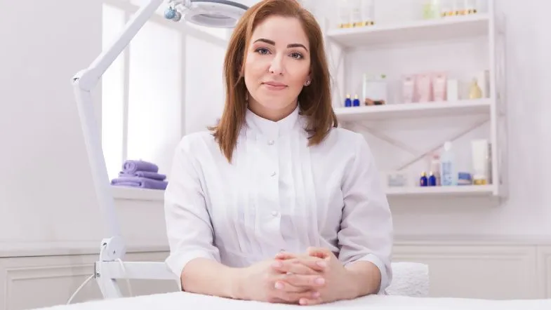 Estética de Alta Performance: formação completa em botox, preenchimento e laserterapia 9 Esteticista ruiva com jaleco branco sentada em ambiente clínico, olhando diretamente para a câmera.