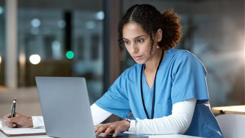 Auditoria hospitalar: como prevenir desperdícios e melhorar indicadores em saúde 10 Profissional de saúde com uniforme azul trabalhando no notebook e anotando em caderno, ilustrando a rotina de auditoria clínica e assistencial.