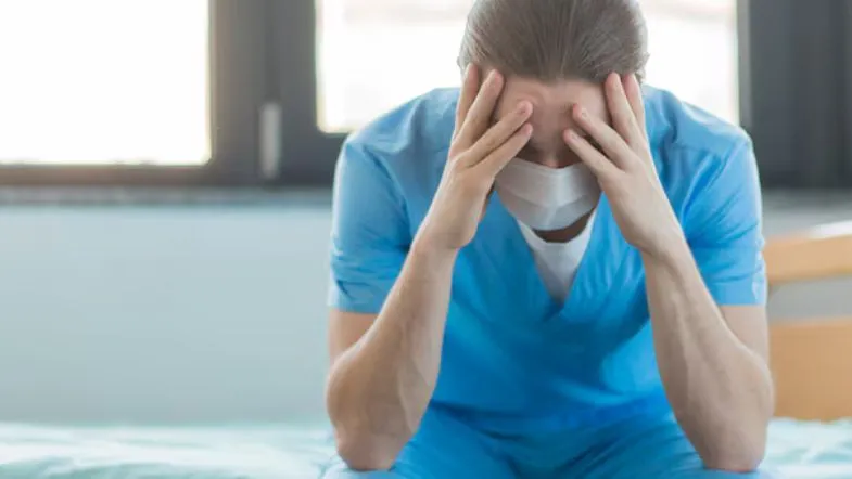 Profissional da saúde sentado com uniforme azul, mãos no rosto, em postura de exaustão emocional.