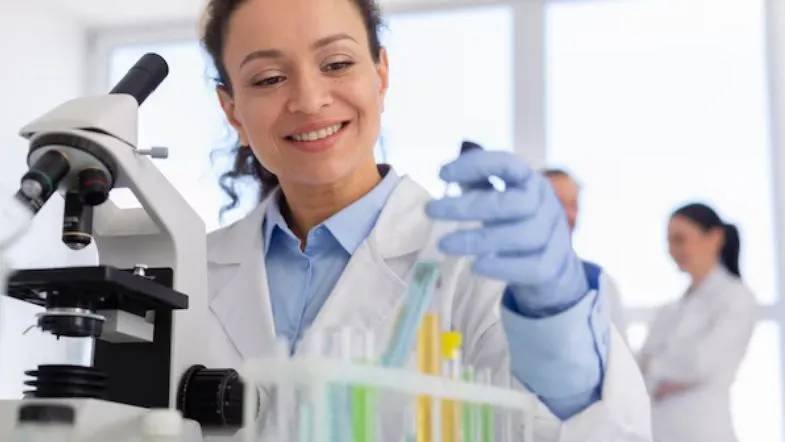 Pesquisadora biomédica manuseando tubos de ensaio ao lado de microscópio.