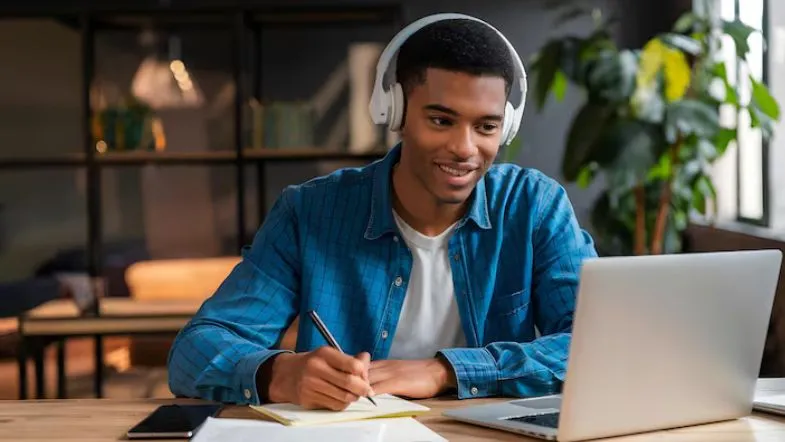 Estudante com fones de ouvido anotando em um caderno enquanto acompanha aula online em um notebook.