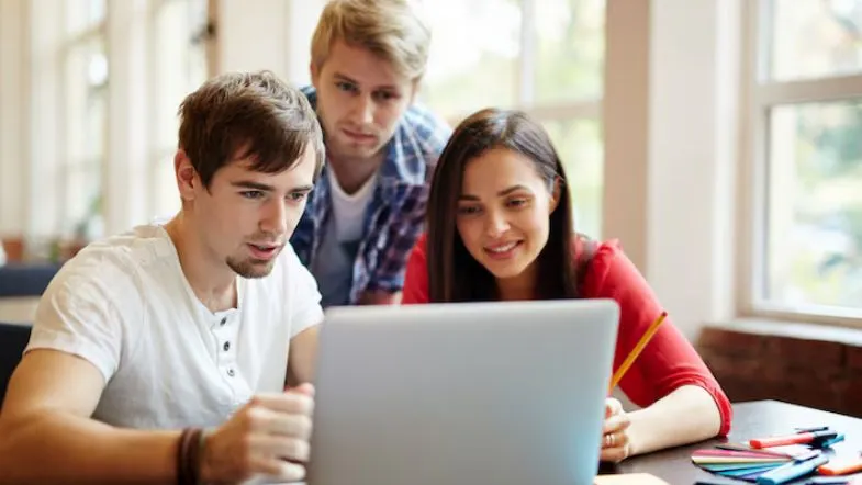 Grupo de três estudantes olhando para um notebook durante estudo em grupo, simbolizando colaboração e aprendizado coletivo.